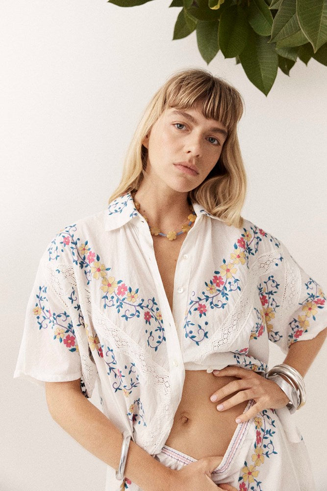 A woman wearing a white short-sleeve button-up shirt with colorful floral embroidery, posed against a white wall with green leaves hanging from above.