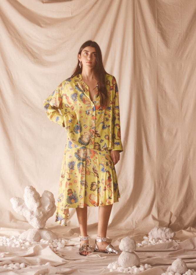 Woman wearing a yellow floral set standing against a beige curtain with decorative elements on the floor.