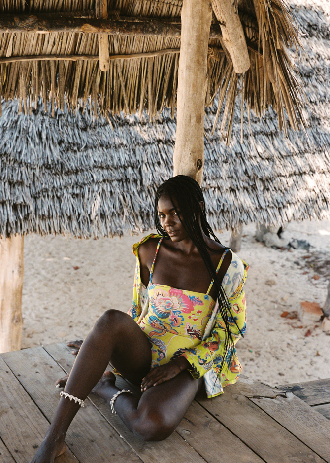 Woman sitting on a wooden platform under a thatched roof structure. Wearing a yellow one-piece swim suit