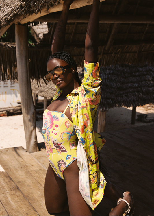 Woman in a yellow swimsuit with a colorful scarf standing under a thatched roof.