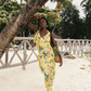 Woman in a yellow floral dress standing under a palm tree on a sandy beach.