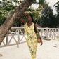 Woman in a yellow floral dress standing under a palm tree on a beach.
