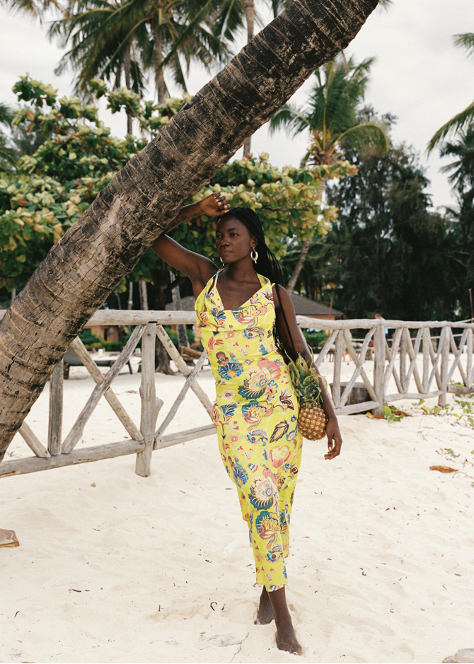 Woman in a yellow floral dress standing under a palm tree on a beach.