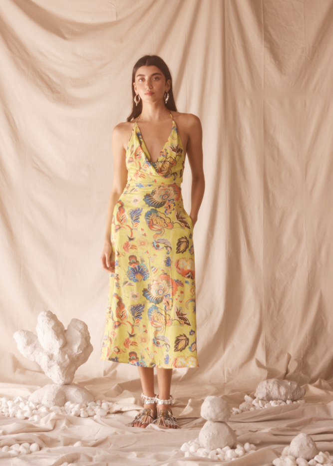 Woman wearing a yellow floral dress standing against a beige curtain with decorative elements on the floor.