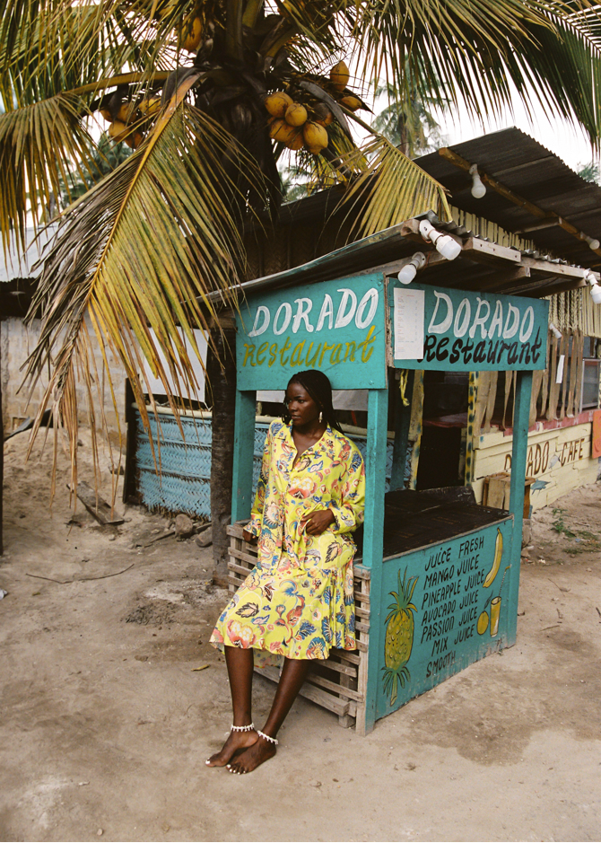 Woman in a yellow floral set sitting in front of a Dorado Restaurant stand with palm trees in the background.