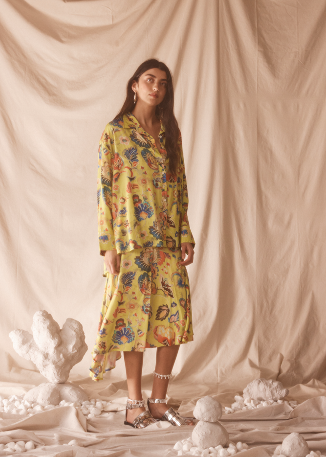 Woman wearing a yellow floral set standing against a beige curtain with decorative elements on the floor.