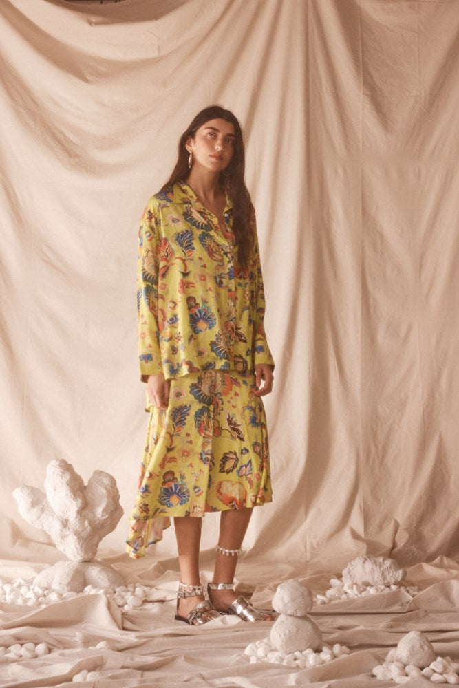 Woman wearing a yellow floral set standing against a beige curtain with decorative elements on the floor.