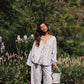 Woman in a light blue shirt and pant set with floral embroided details standing in a garden with greenery and flowers.