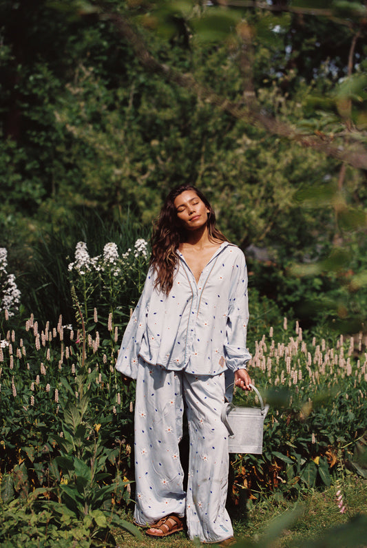 Woman in a light blue shirt and pant set with floral embroided details standing in a garden with greenery and flowers.