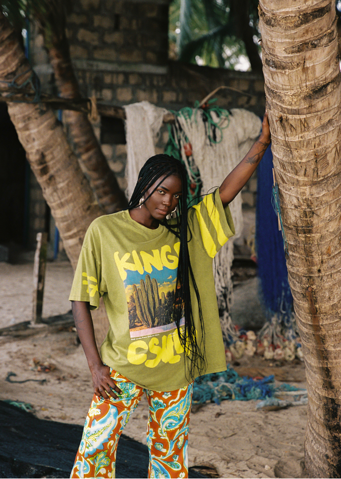 Person wearing a lime green t-shirt with desert text and colorful pants standing next to a palm tree.