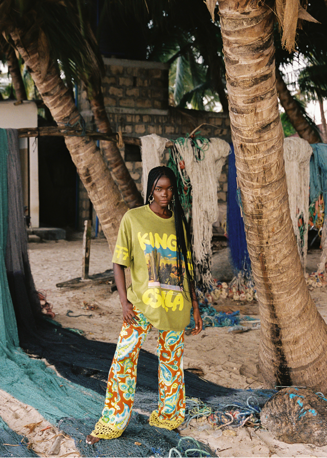 Person wearing a bright lime green shirt with desert text and colorful pants standing under palm trees.