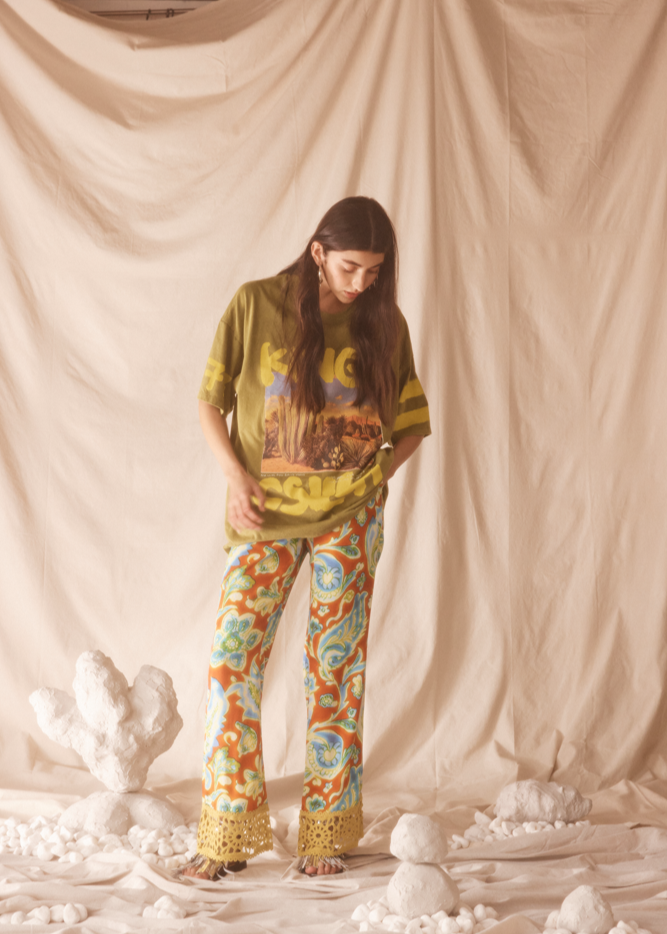 Person wearing a lime green patterned shirt and colorful pants standing against a beige curtain with decorative elements.