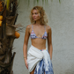 Woman in a pink patterned bikini and sarong standing next to a palm tree with fruit.