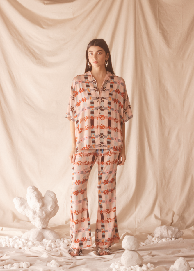 Woman wearing a pink patterned outfit standing against a beige curtain with decorative elements on the floor.