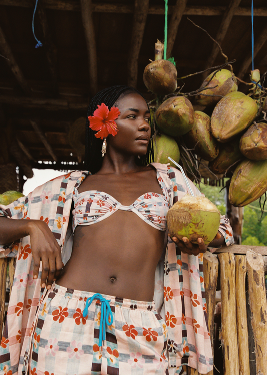 Woman in a tropical setting holding coconuts with hanging coconuts in the background wearing pink floral set