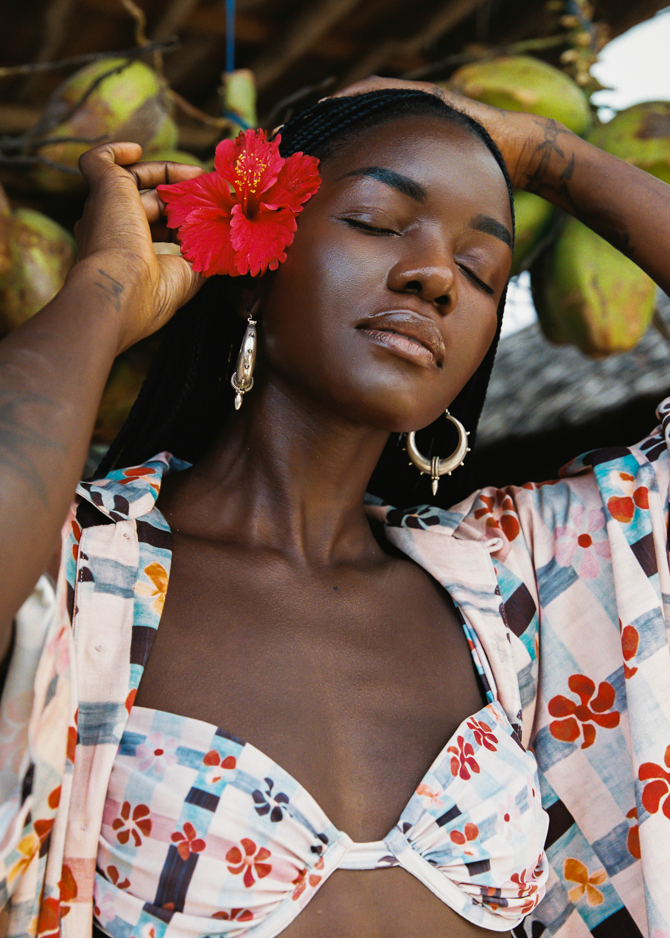 Woman with a red flower in her hair, wearing a colorful pink bikini and patterned cover-up, surrounded by coconuts.