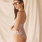 Woman wearing a pink floral one-piece swimsuit and sunglasses against a beige curtain background