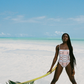 Woman in a pink patterned swimsuit standing on a sandy beach with clear blue water and sky.