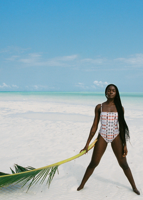 Woman in a pink patterned swimsuit standing on a sandy beach with clear blue water and sky.