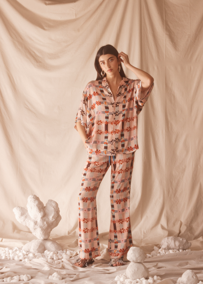 Woman wearing a pink patterned shirt and flare pants standing against a beige curtain with decorative elements.