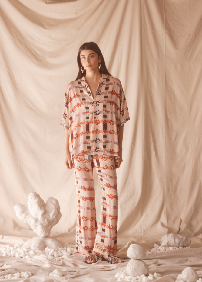 Woman wearing a pink floral patterned outfit standing against a beige curtain with decorative elements on the floor.