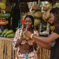Two people interacting with coconuts at a market stall. Women is wearing pink patterned matching set and bikini top.
