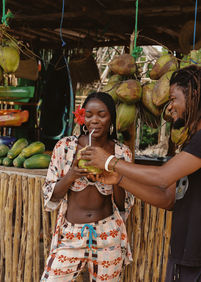 Two people interacting with coconuts at a market stall. Women is wearing pink patterned matching set and bikini top.
