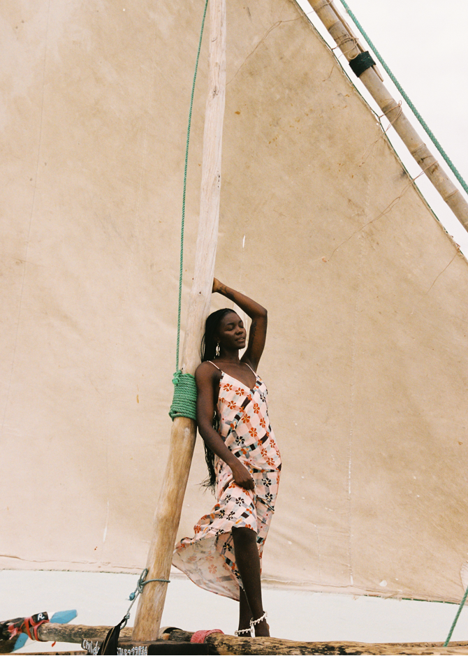 Woman in a pink floral dress standing on a traditional wooden boat with a large beige sail.