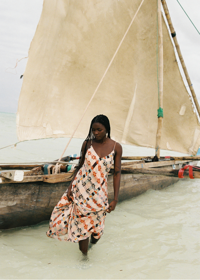Woman in a pink floral dress standing in shallow water near a traditional sailboat.