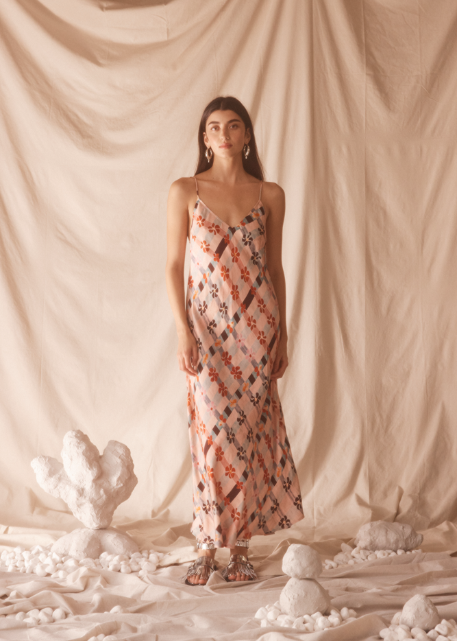 Woman in a pink floral dress standing against a beige curtain with decorative elements on the floor.