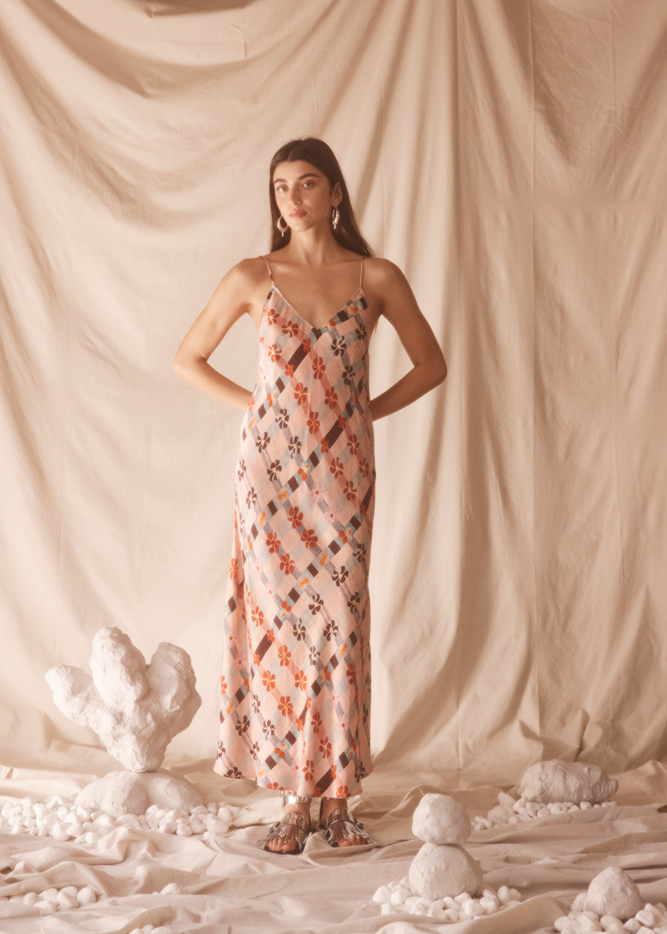 Woman in a pink floral dress standing against a beige curtain with decorative elements on the floor.