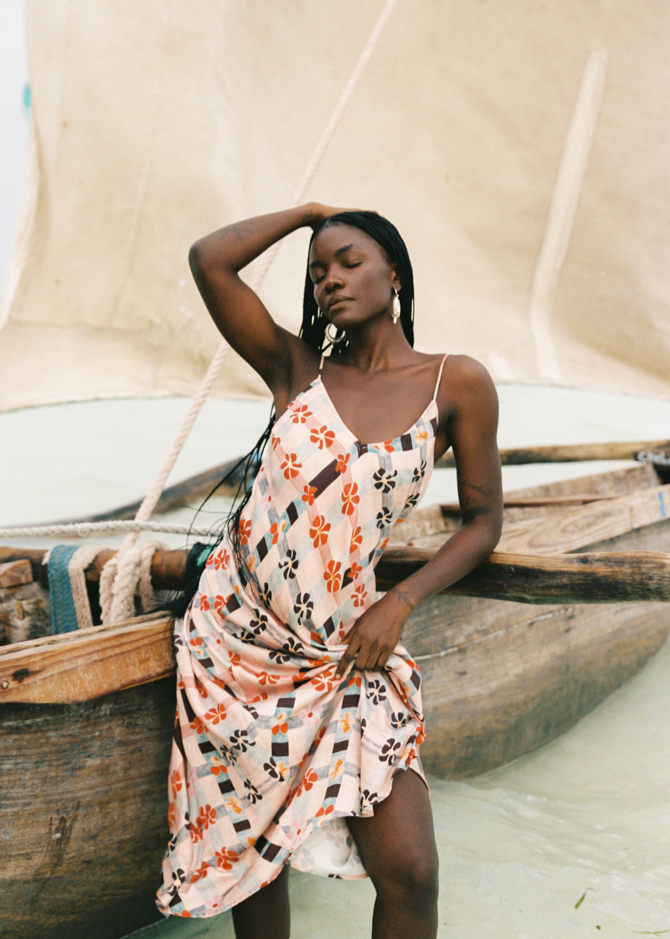 Woman in a pink floral dress standing next to a wooden boat with a sail in the background