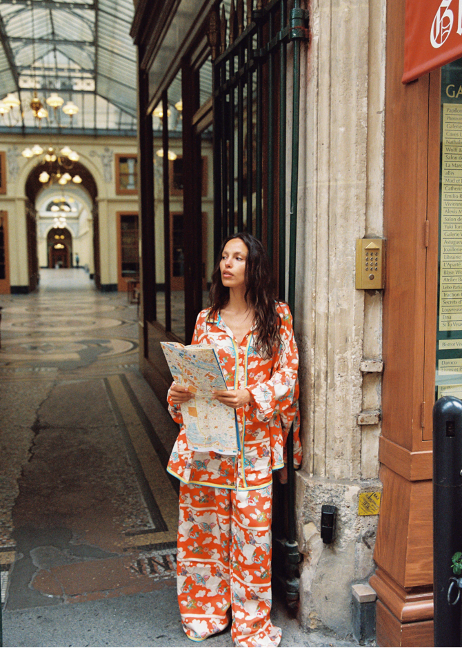 Woman in an orange patterned outfit with cloud/butterfly print standing in an architectural hallway, holding a map.