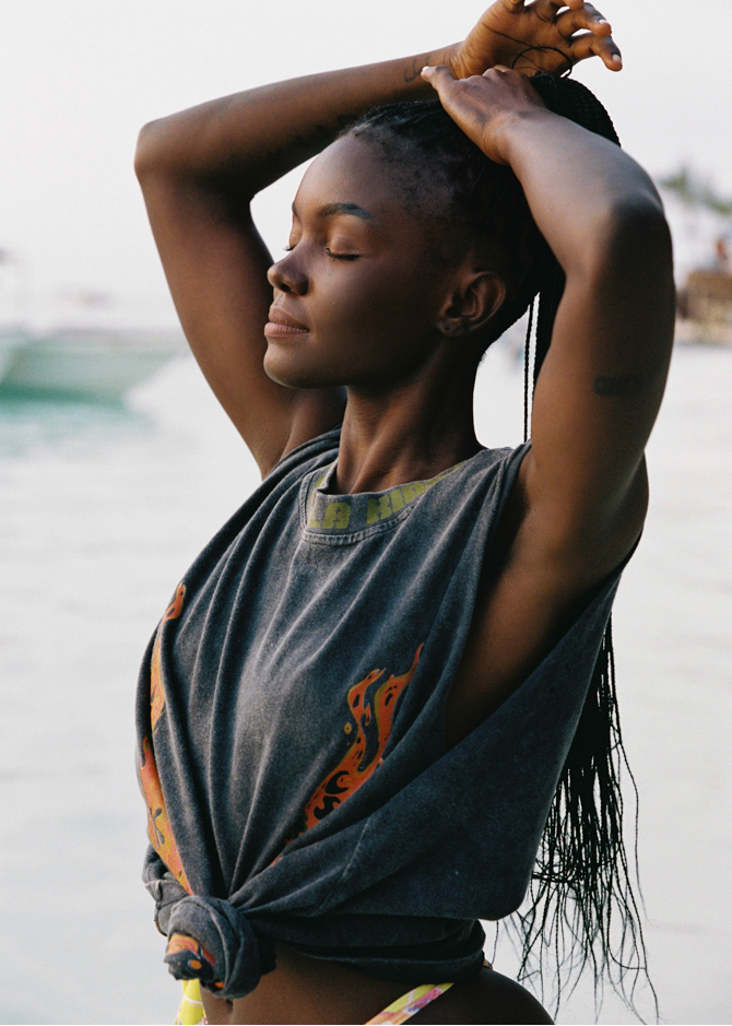 Woman with braided hair wearing a dark gray tank with colorful patterns, standing by water.