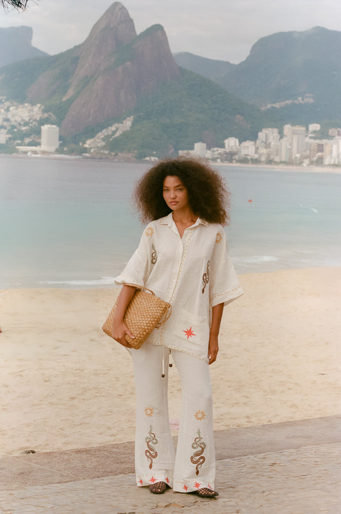 Woman in a white shirt and pant outfit with snake patterns standing on a beach with mountains in the background