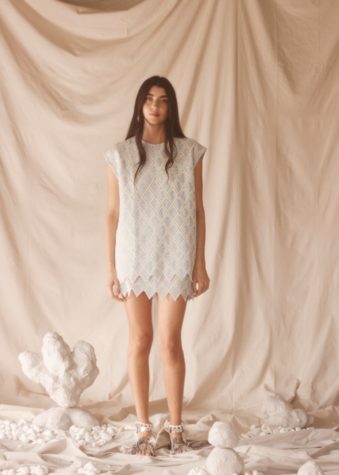 Woman wearing a light blue embroided set standing against a beige curtain with decorative elements.