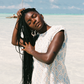 Woman with long braided hair standing on a sandy beach with clear blue sky wearing a light blue blouse