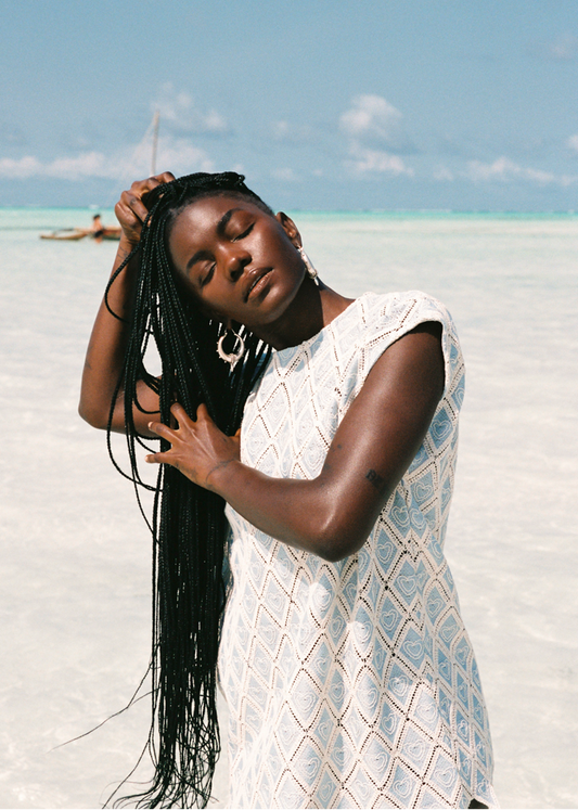 Woman with long braided hair standing on a sandy beach with clear blue sky wearing a light blue blouse