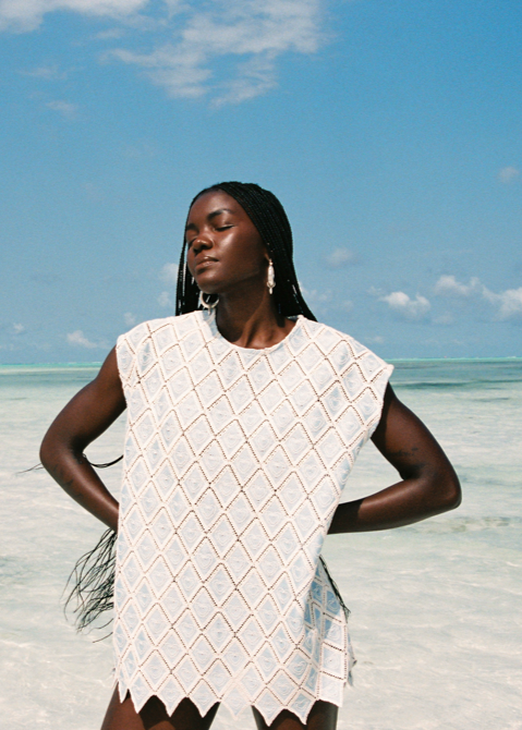 Woman wearing a light artisional blue set on a beach with clear blue sky and water.