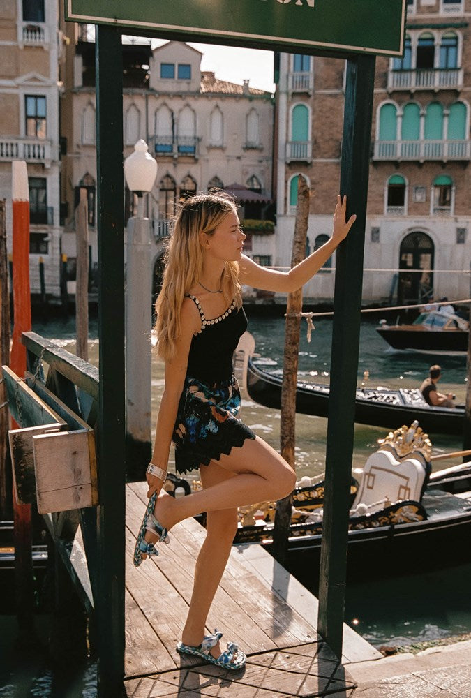 A woman posing on a wooden dock in Venice, wearing a black sleeveless dress with a colorful floral patterned hem and blue patterned shoes, with gondolas and historic buildings in the background.