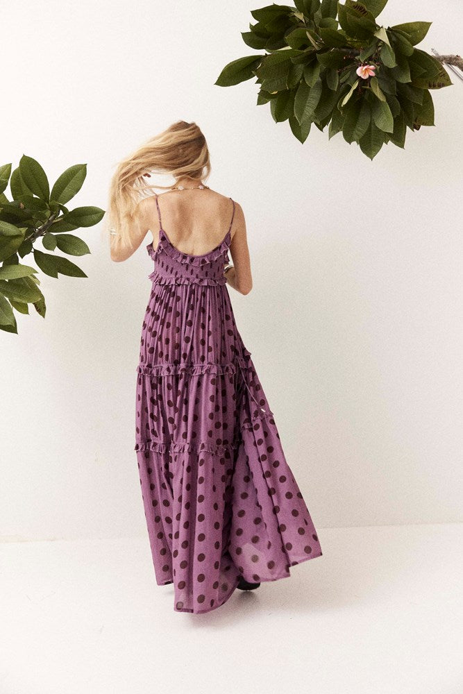 A woman wearing a purple tiered maxi dress with dark brown polka dots, shown from the back against a white wall with green plants in the corners.