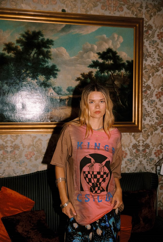 A woman stands in a vintage room wearing a faded pink graphic t-shirt with 'KINGA CSILLA' printed on the front, paired with blue and black patterned pants. Behind her is a large framed landscape painting and floral wallpaper.