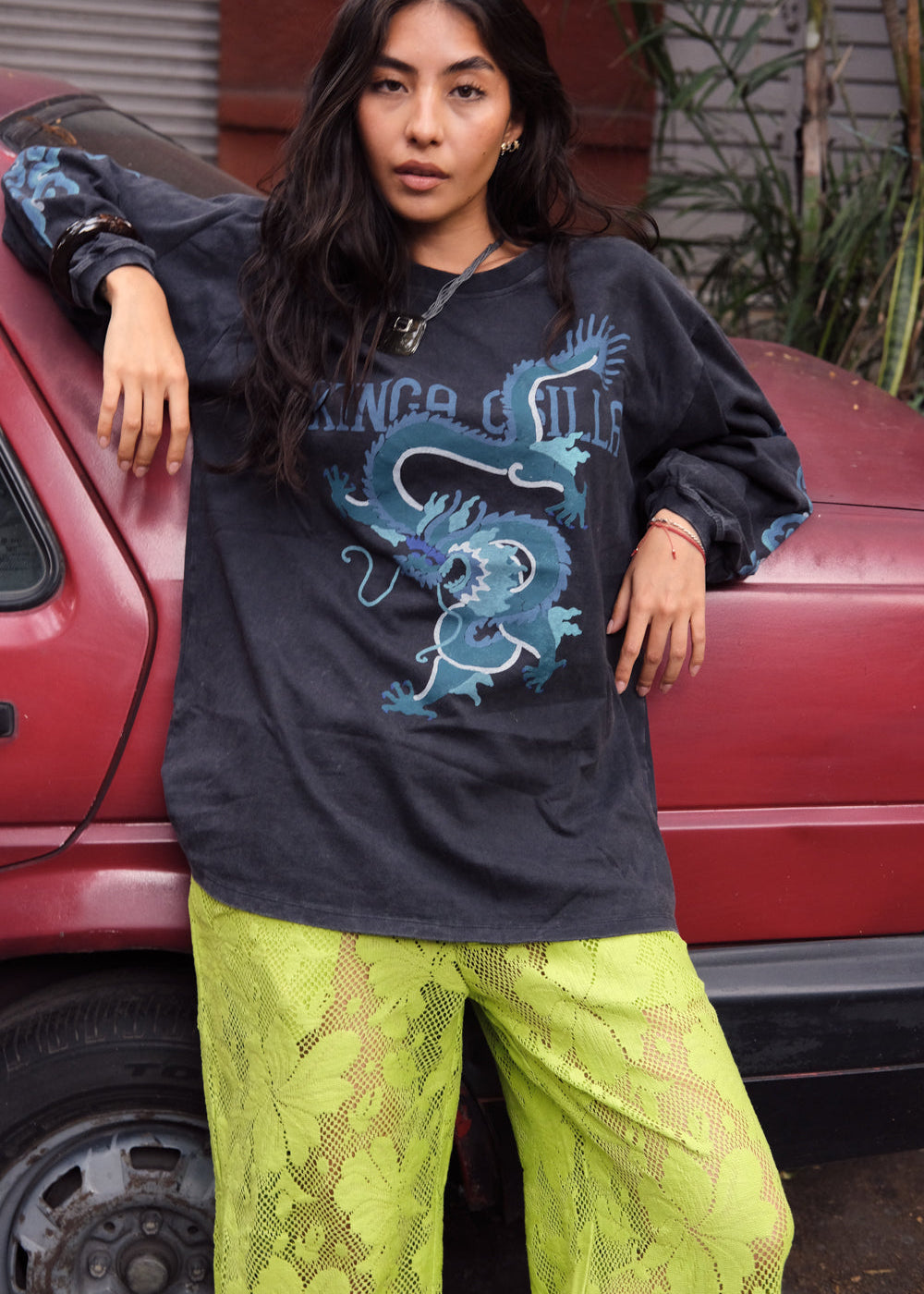 Person wearing a black sweatshirt with a blue dragon design and green pants, leaning against a red vehicle.