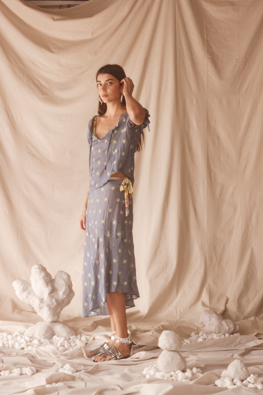 Woman in a sheer, blue blouse and midi skirt set with yellow sun embroided detailing.