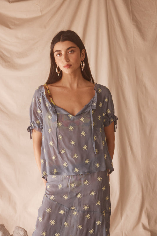 Woman wearing sheer blue blouse with delicate sunburst embroidery, drawstring neckline, and airy cropped silhouette for summer style.
