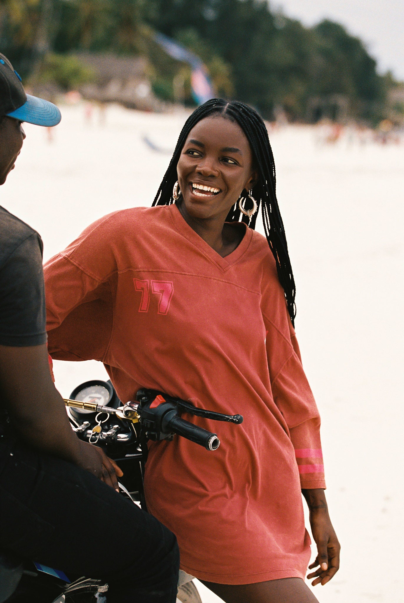 Woman in a red shirt dress standing next to a motorcycle on a beach.