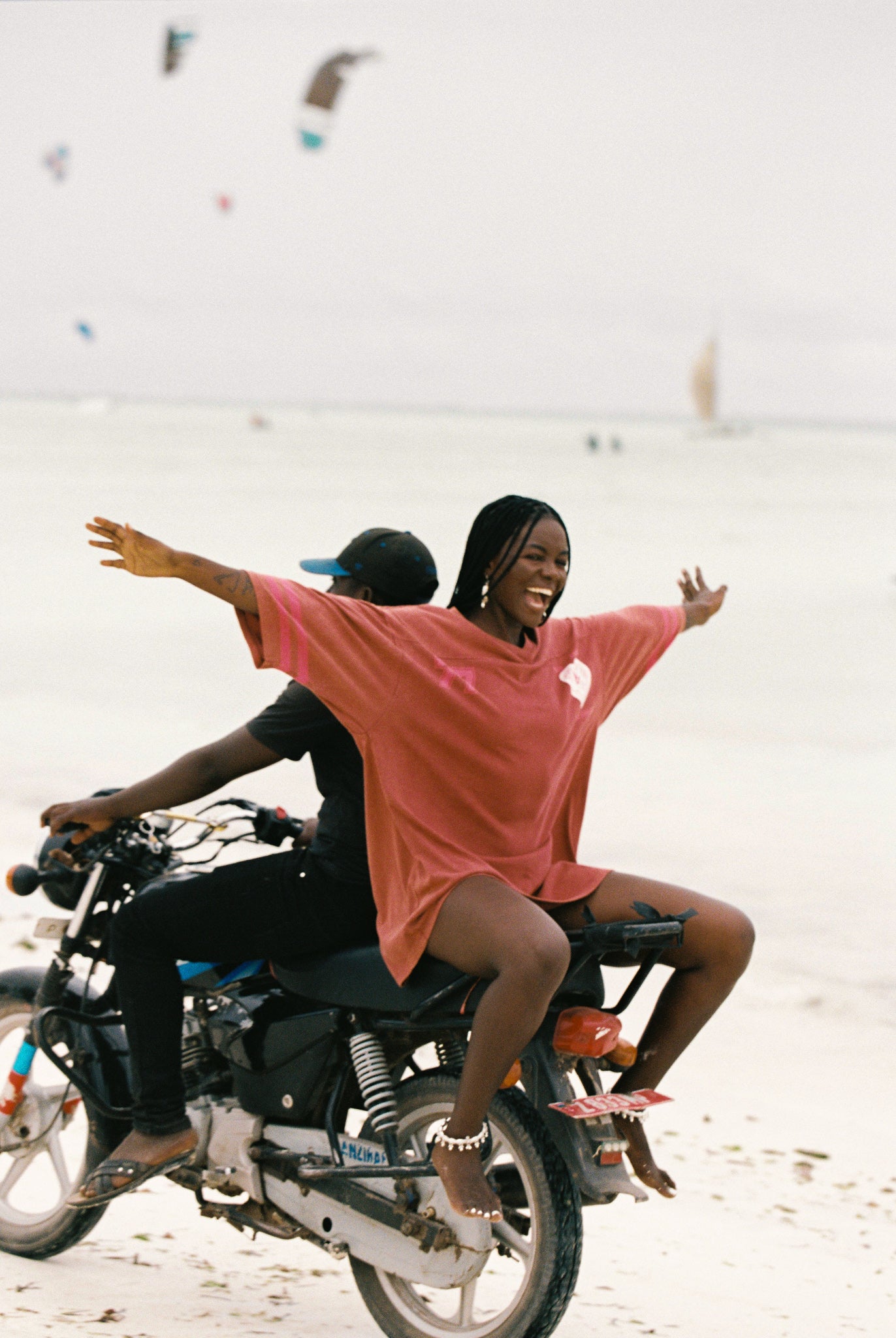 Two people on a motorcycle with one person in a red t-shirt dress, riding on a beach.