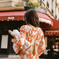 Back view of model wearing the Kinga Csilla Farfalla Olive Dress, a long-sleeve orange maxi with butterfly and floral print, carrying fresh baguettes outside a Paris café with red awning.