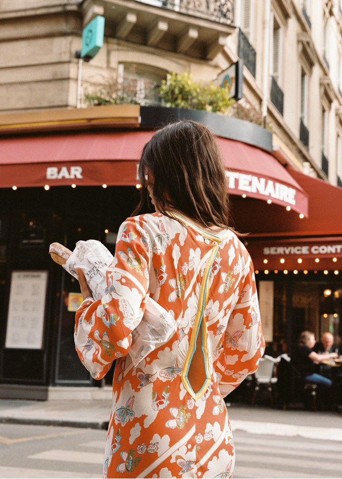 Back view of model wearing the Kinga Csilla Farfalla Olive Dress, a long-sleeve orange maxi with butterfly and floral print, carrying fresh baguettes outside a Paris café with red awning.