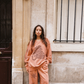 Model leaning against a stone wall in Paris wearing Kinga Csilla Abracadabra Hades Pant with matching Abracadabra OG Jumper in burnt orange with sun motif